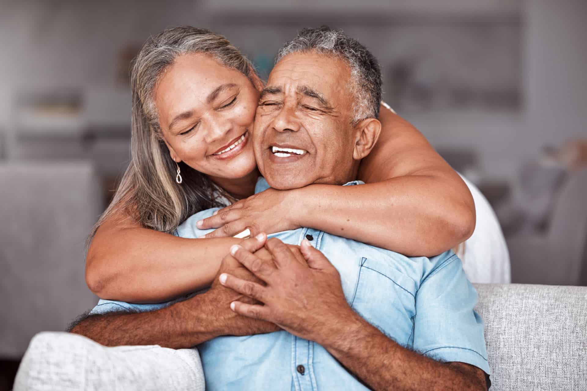 Happy Senior Couple, Hug And Relax In Love For Relationship Bonding Together In Tender Happiness At Home. Joyful Elderly Man And Woman Smile In Hope Embracing Romance For Retirement House On Sofa